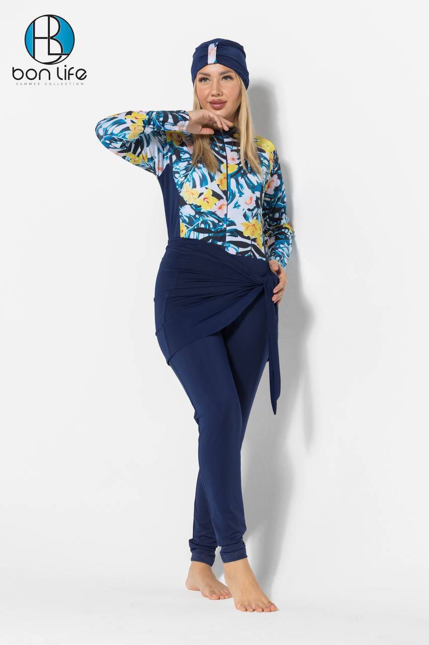 Woman wearing a colorful floral long-sleeve top and navy pants with a matching cap, standing against a white background.